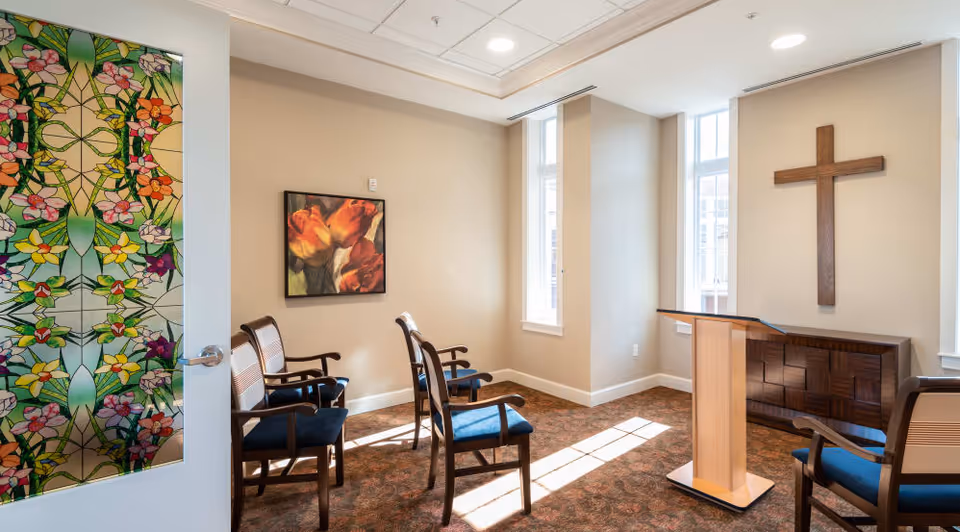 A small chapel or prayer room with a wooden cross mounted on the wall, a wooden podium, and several wooden chairs with blue cushions arranged facing the podium. The room has beige walls, carpeted floor, and two tall windows letting in natural light. The door has a colorful stained glass design with floral patterns.