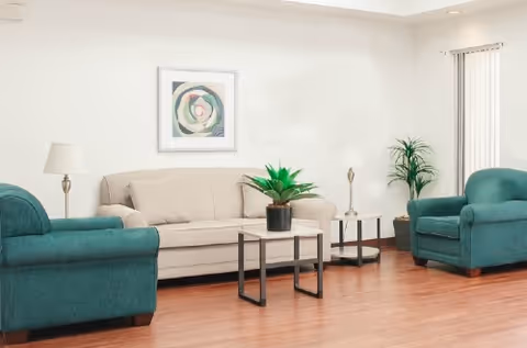 A living room with a beige sofa and two teal armchairs arranged around a small white coffee table with a potted plant. There are two floor lamps on either side of the sofa, a framed abstract artwork on the wall, and a tall potted plant near a window with vertical blinds.