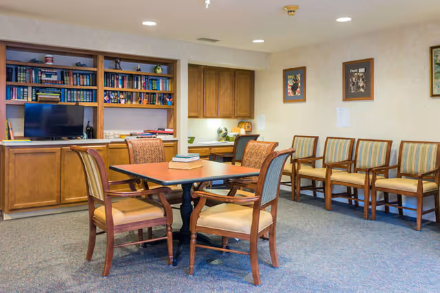 A cozy common area with a square wooden table surrounded by four cushioned chairs. Behind the table, there is a built-in wooden bookshelf filled with books and decorative items, along with a small television. To the right, there are several wooden chairs with striped cushions lined up against the wall, which is decorated with framed pictures. The room has a carpeted floor and recessed ceiling lights.