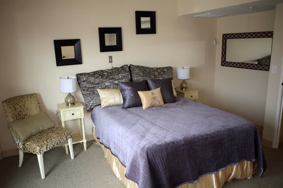 A bedroom with a bed covered in a purple quilt and multiple decorative pillows. There are two small nightstands on either side of the bed, each with a lamp. A patterned chair with a matching cushion is placed next to one nightstand. Three square mirrors are hung on the wall above the bed, and a larger rectangular mirror is on the adjacent wall.