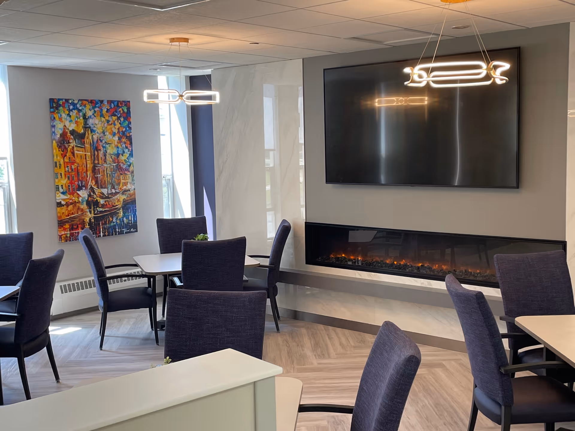 A modern dining area with several tables and dark upholstered chairs. There is a large flat-screen TV mounted above a long electric fireplace on the wall. A colorful painting hangs on the wall near two windows letting in natural light. Contemporary pendant lights hang from the ceiling.