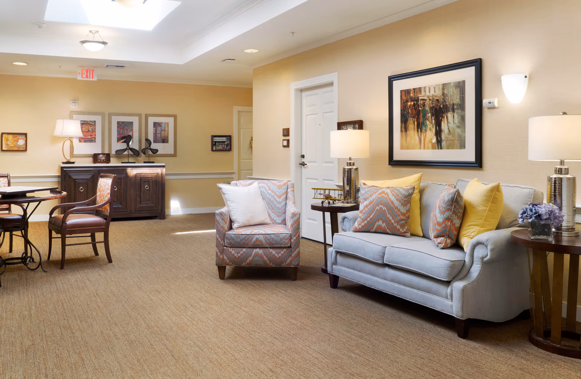 A cozy living room area in a senior living facility with a light blue sofa adorned with colorful pillows, a matching armchair with a white pillow, two side tables with lamps, a framed painting on the wall, and a wooden cabinet with decorative items. The room has beige walls and carpeted flooring.