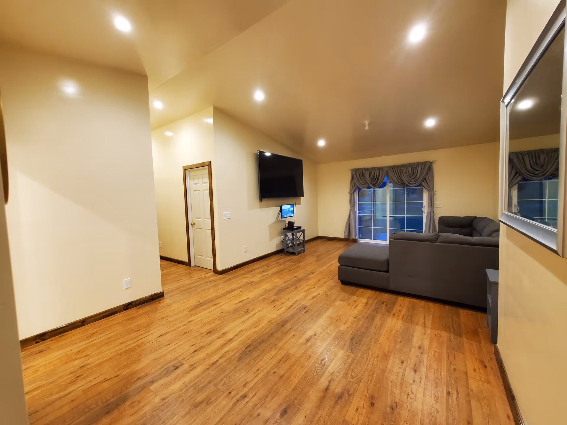 A spacious living room with wooden flooring, beige walls, and recessed ceiling lights. There is a gray sectional sofa near a sliding glass door with curtains, a wall-mounted flat-screen TV, and a small stand beneath the TV. A large mirror is mounted on the right wall.