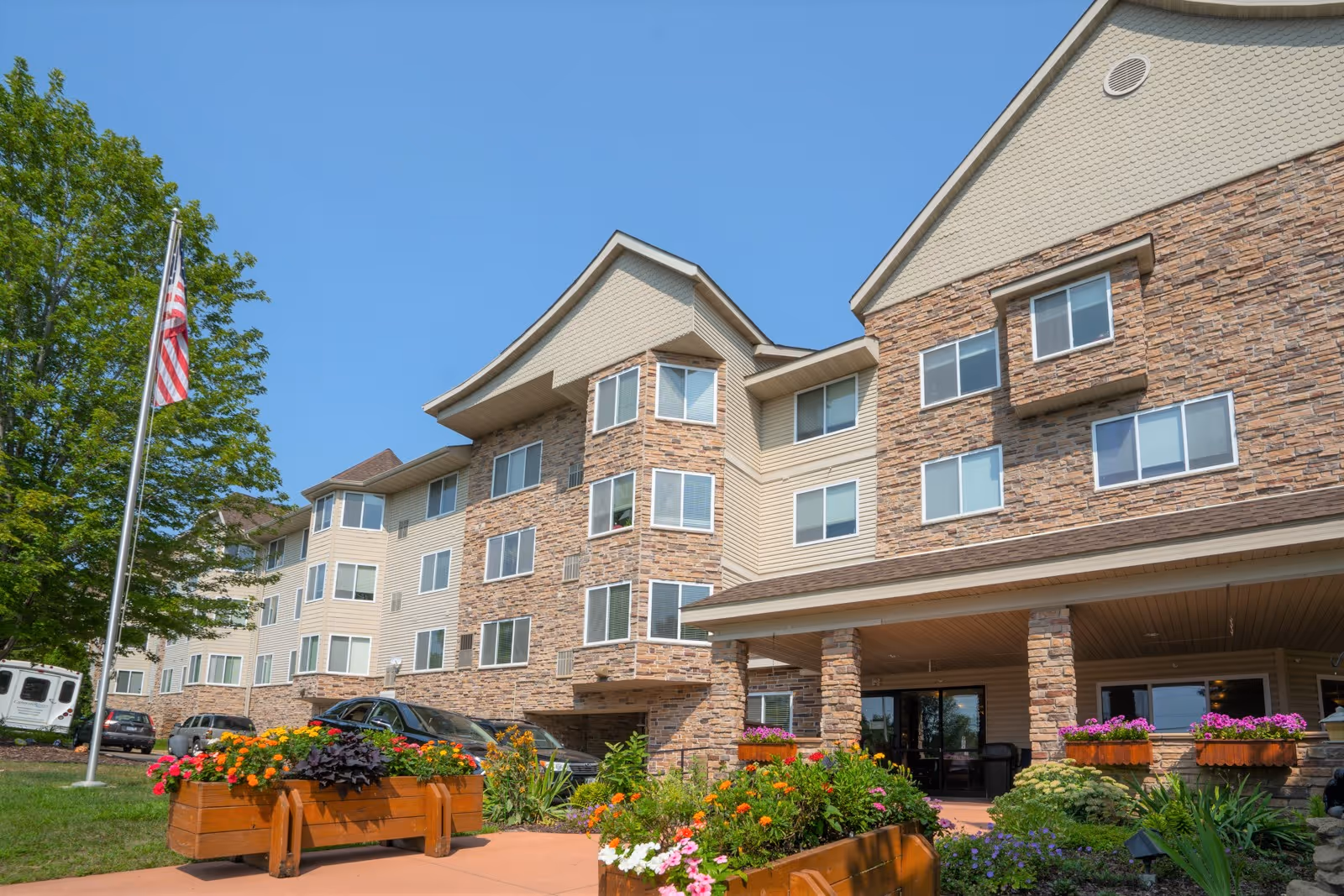 Exterior view of Cannon Rivers Senior Living building with a stone and beige siding facade, multiple windows, and a covered entrance. There are colorful flower planters in the foreground, a flagpole with an American flag, and several parked cars visible.