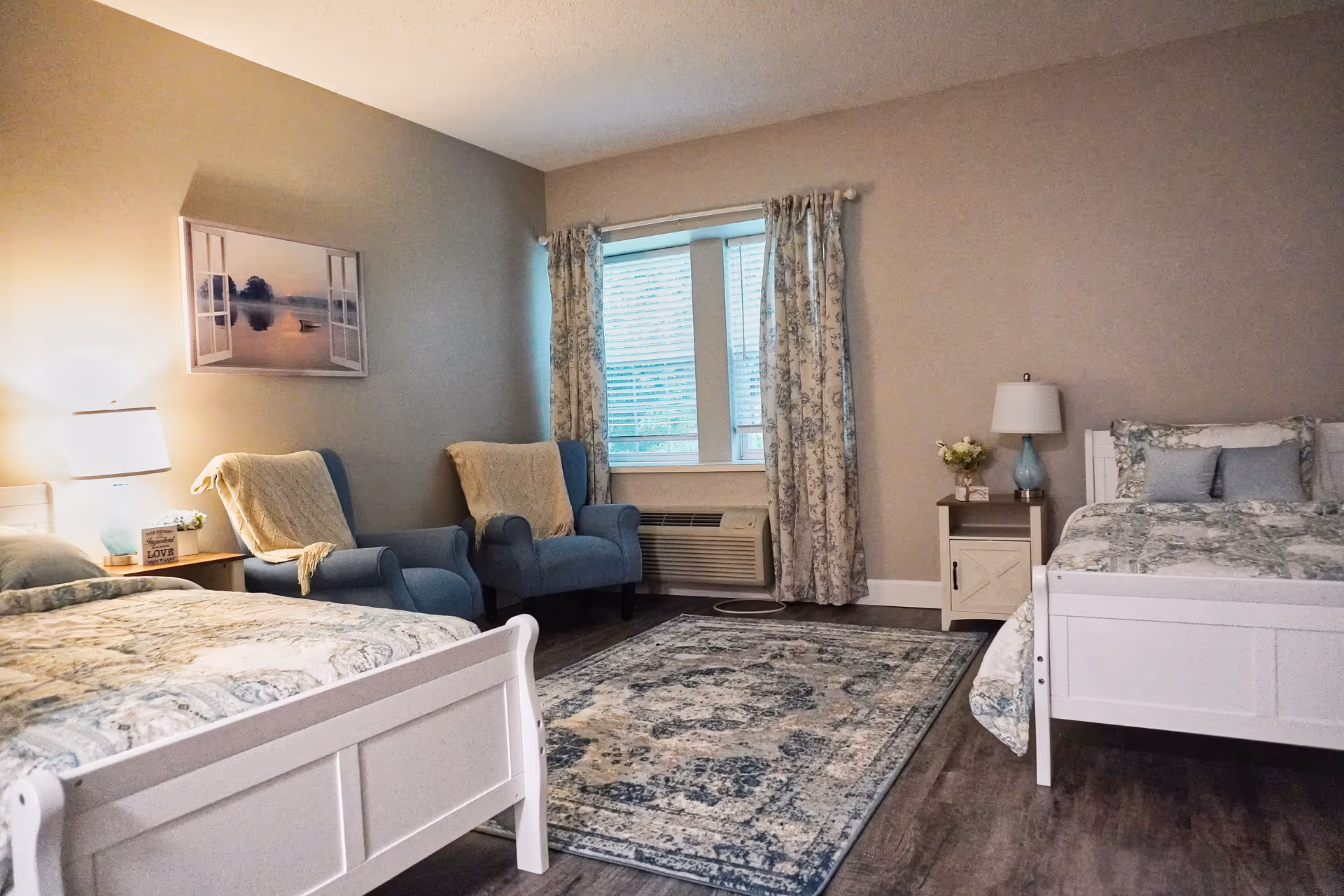A cozy bedroom in an assisted living facility featuring two white wooden beds with patterned bedding, two blue armchairs with beige throws, a window with floral curtains, a patterned area rug, and bedside tables with lamps and decorative items.