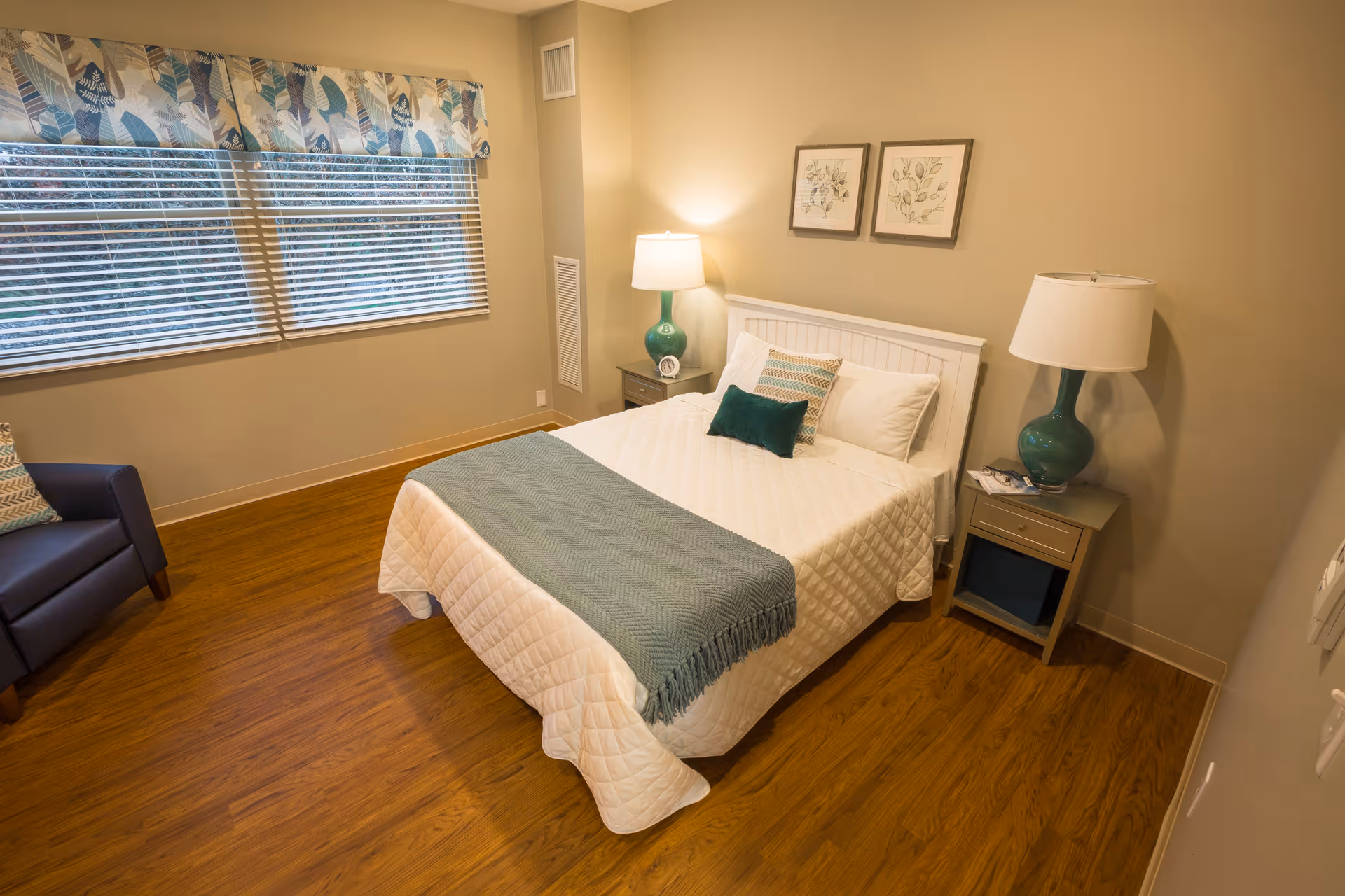 A cozy bedroom with a neatly made bed featuring white bedding and teal accent pillows and throw. There are two matching bedside tables with teal lamps, a window with blinds and a floral valance, a dark armchair in the corner, and two framed botanical prints on the wall above the bed. The floor is wooden and the walls are painted beige.