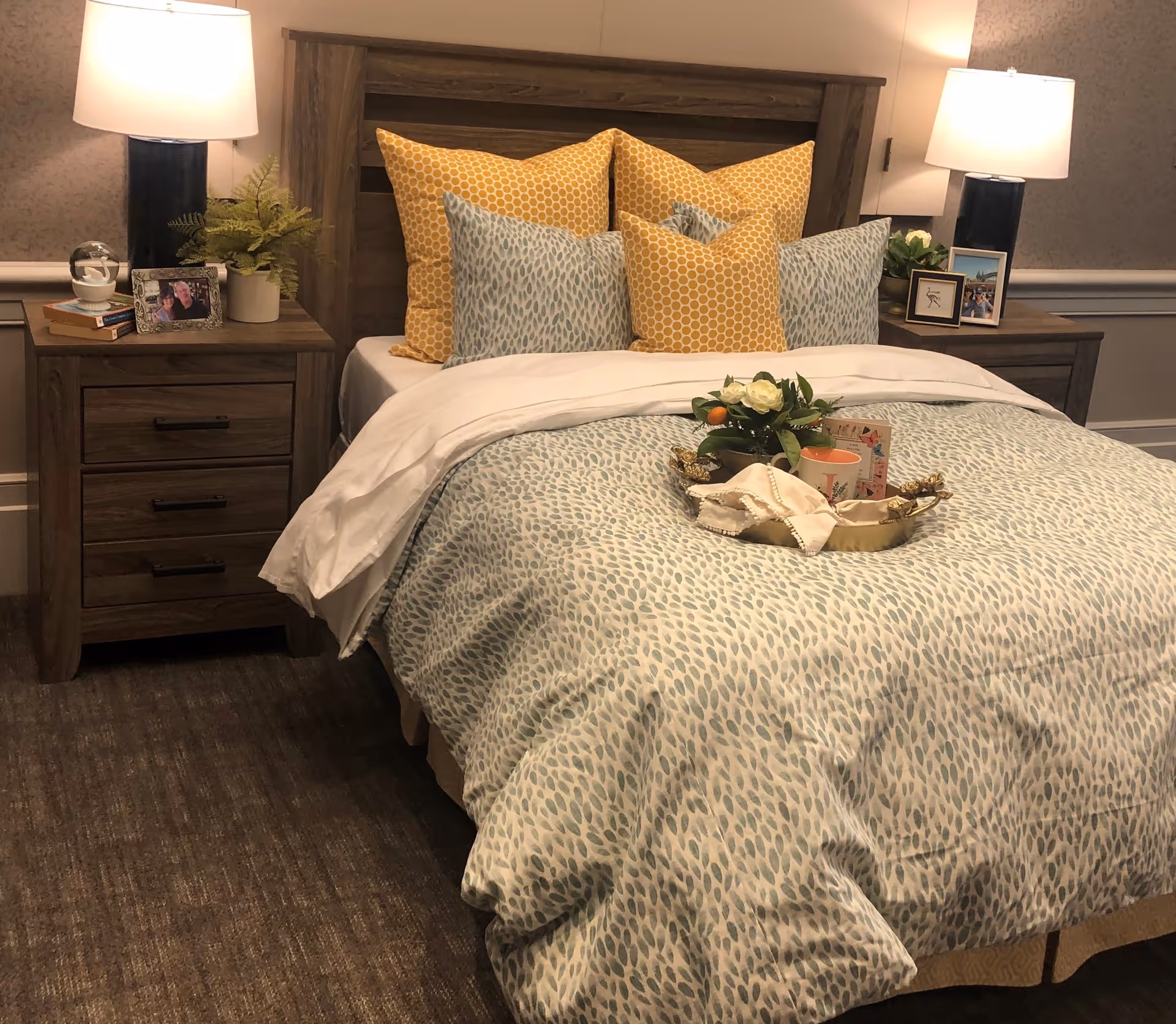 A neatly made bed with a wooden headboard, decorated with multiple yellow and blue patterned pillows. There are two wooden nightstands on either side of the bed, each with a lamp, framed photos, and small plants. A tray with a flower arrangement and a cup is placed on the bed. The room has a carpeted floor and neutral-colored walls.