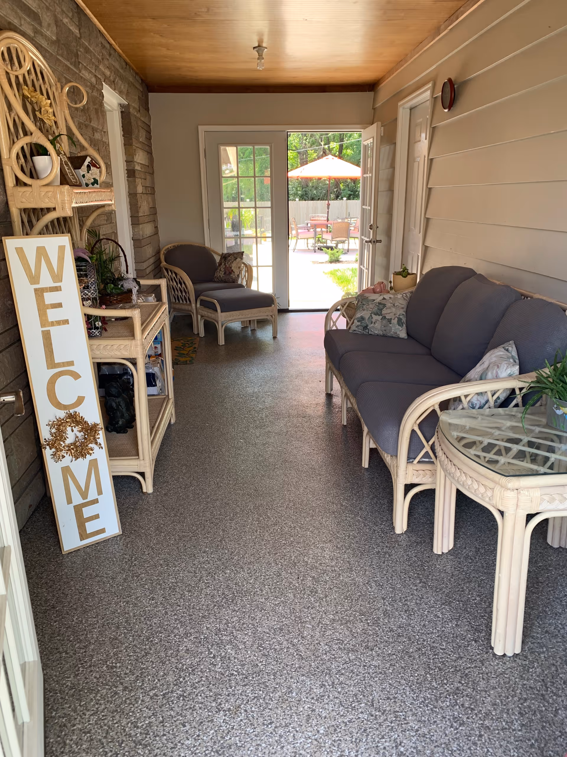 A cozy indoor seating area with wicker furniture including a sofa, armchair, and glass-top side table. A tall welcome sign leans against the wall on the left, and there are plants and decorative items on shelves. French doors at the far end open to an outdoor patio with a table and chairs under a red umbrella.