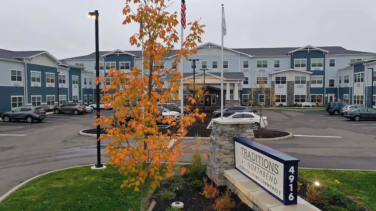 Front exterior of the Traditions at North Bend senior living building with a parking lot, fall-colored tree, and entrance sign.