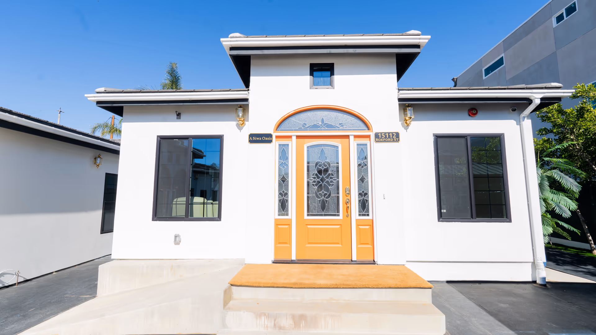 Small white building front with an orange decorative entrance door flanked by two windows under a clear blue sky.