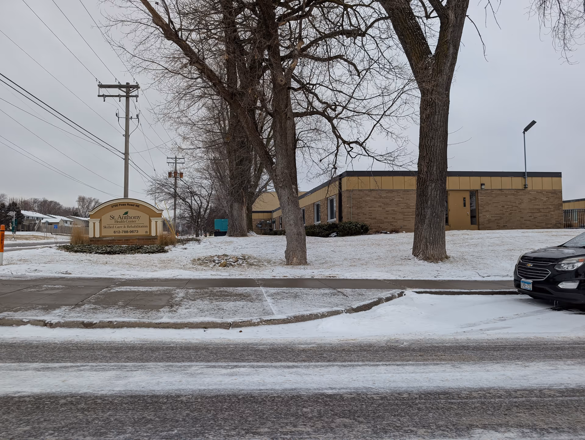 Snowy exterior view of the St. Anthony Health & Rehabilitation building with a stone sign, leafless trees, and a parked car.