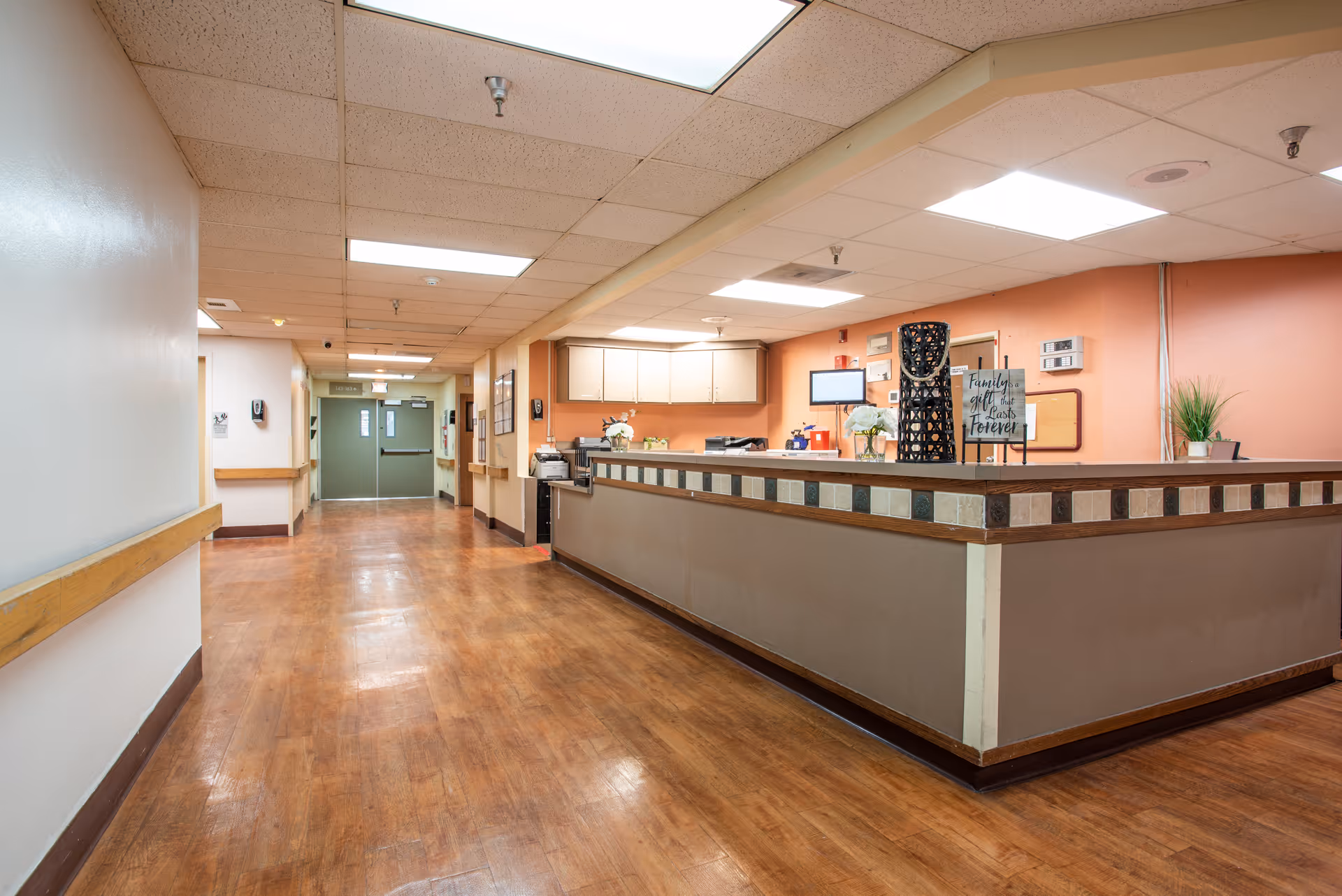 Interior hallway with a reception/nurse station desk on the right, wood-look floors, and overhead fluorescent lighting.