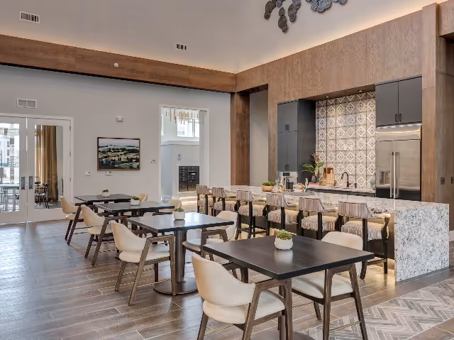 A modern dining area with several dark wood tables and beige cushioned chairs. In the background, there is a kitchen area with a marble countertop, bar stools, and a tiled backsplash. The room has high ceilings and large windows allowing natural light to enter.