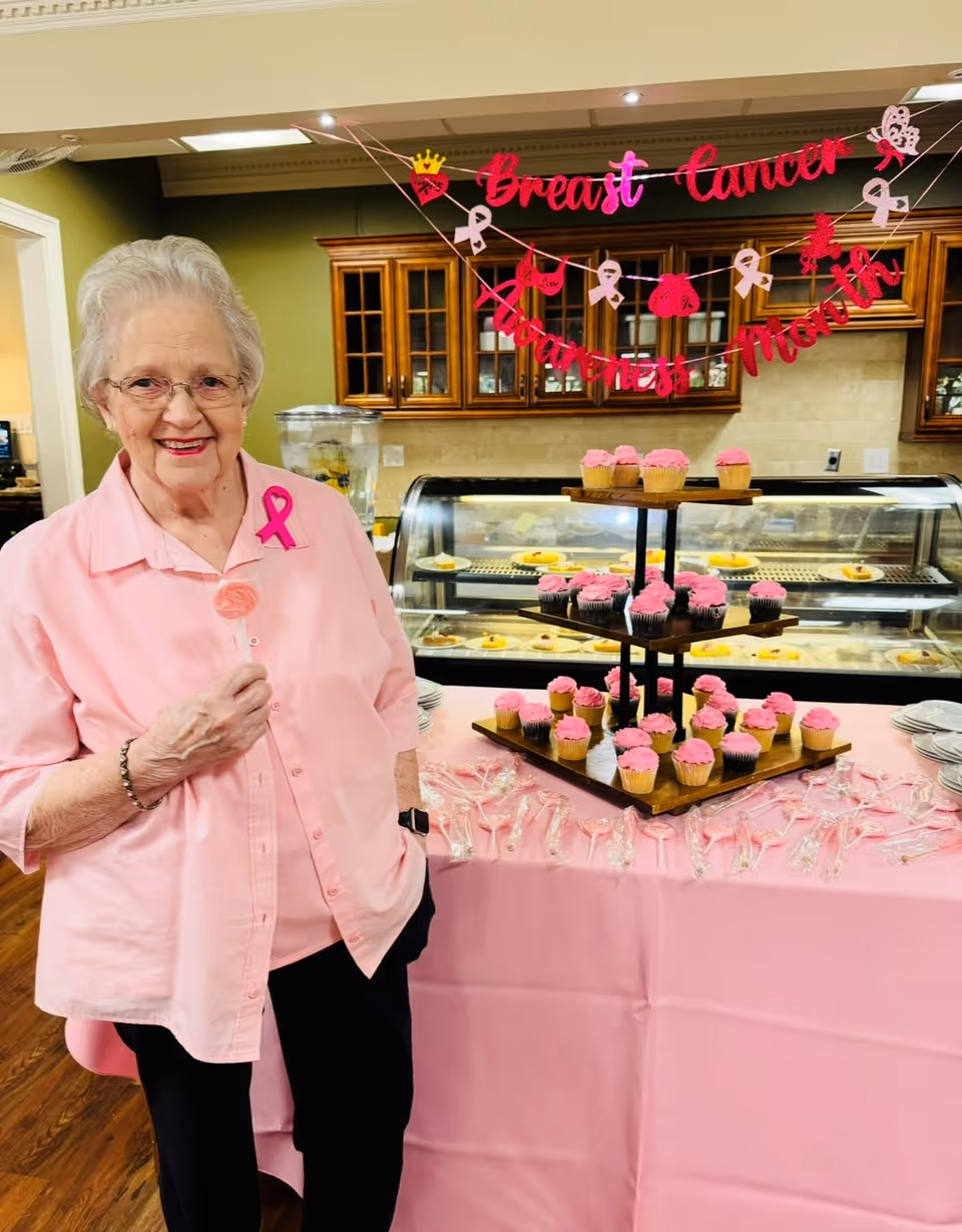 An elderly woman in a pink shirt stands beside a table of pink-frosted cupcakes under a 'Breast Cancer Awareness' banner in a dining area.