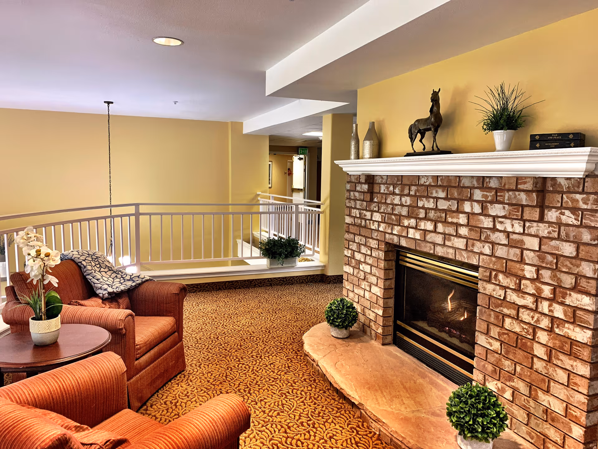 Cozy senior living common area with two red armchairs, a round wooden table with a white orchid plant, a brick fireplace with a small fire burning, decorative plants, and a horse statue on the mantel. The walls are painted yellow and the carpet has a patterned design.
