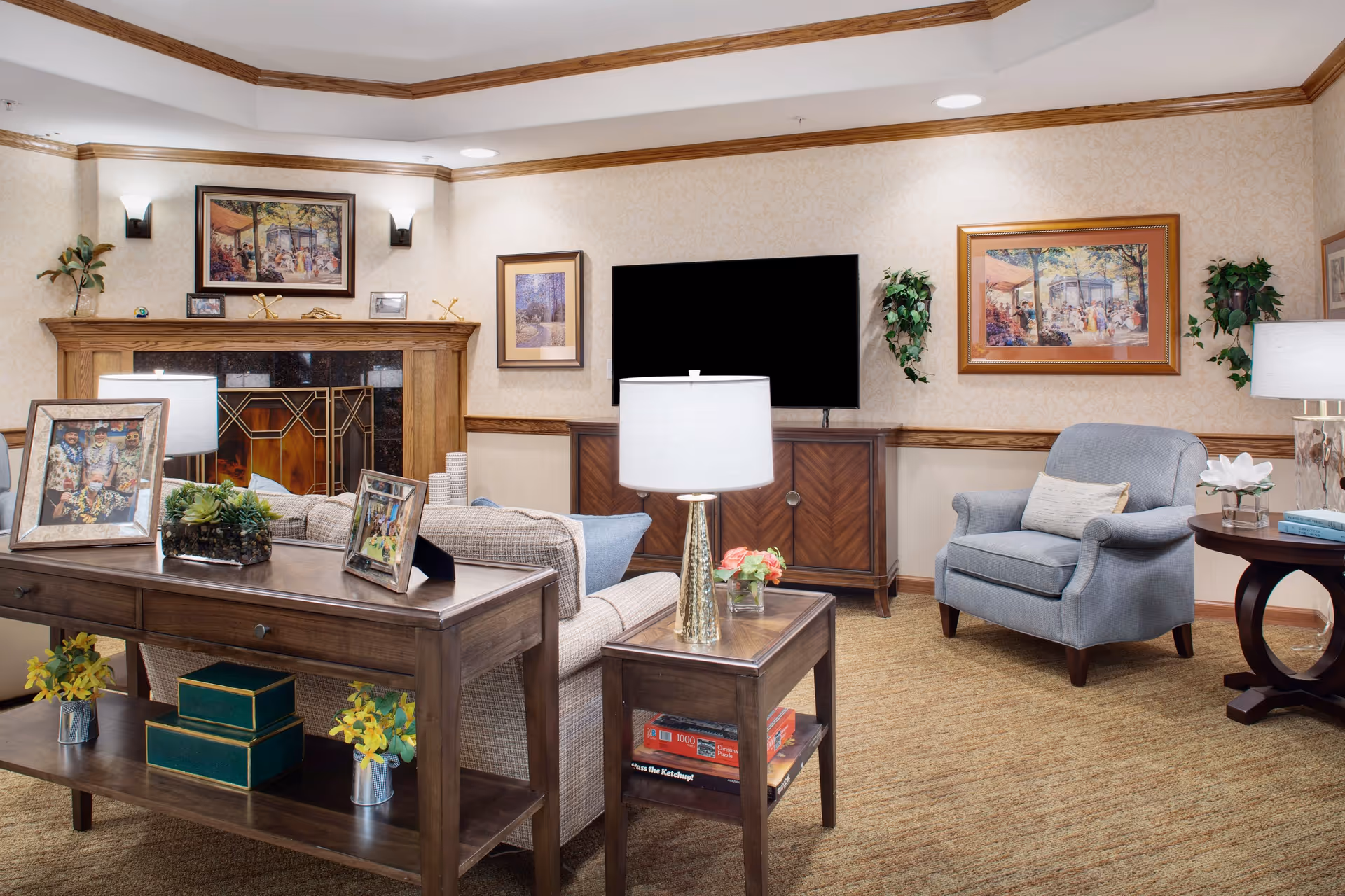 A cozy living room with a beige sofa, a blue armchair, and wooden furniture including a console table and side table. The room features a fireplace with a decorative screen, a flat-screen TV on a wooden cabinet, framed artwork on the walls, table lamps, and small plants and flowers as decor.