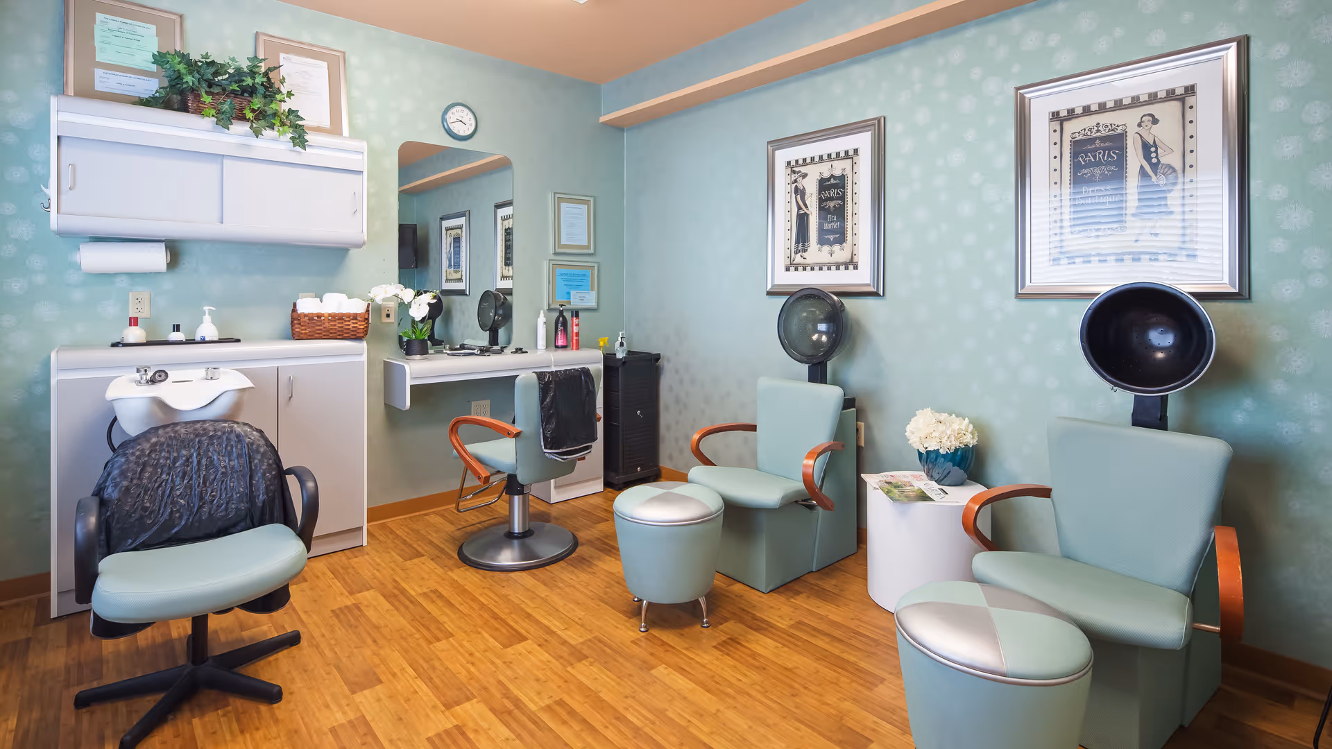 Interior of a hair salon room with light green walls and wooden floor. The room contains salon chairs, a hair washing station, two hair dryers, a mirror, framed artwork on the walls, and a small table with a flower vase and magazines.