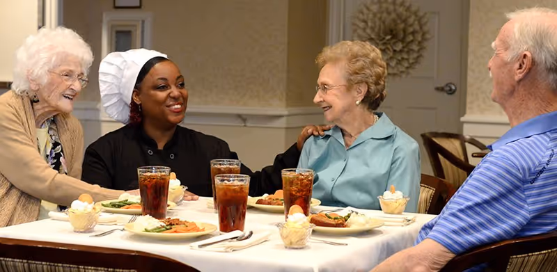A chef and three elderly residents smile and chat around a dining table set with plates of food and glasses of iced tea.