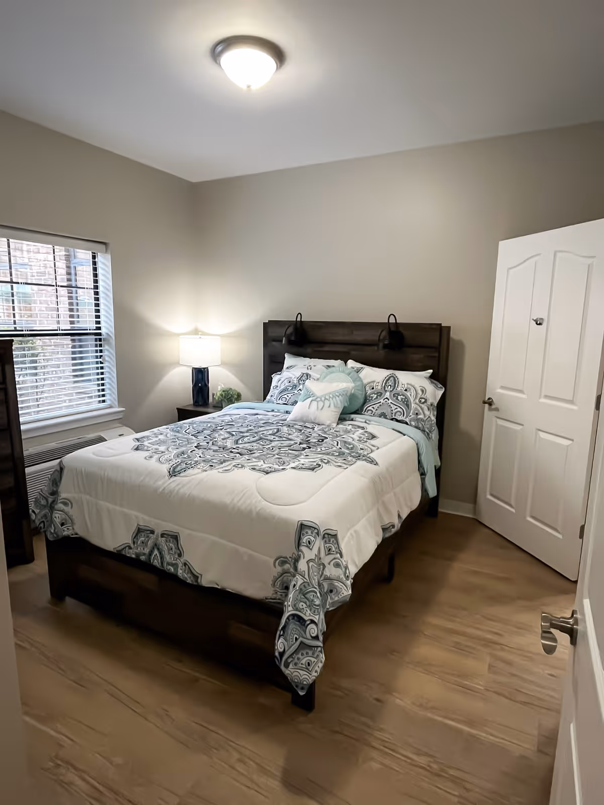Neatly made bed with patterned bedding in a softly lit bedroom with a nightstand, lamp, window, and closed door.