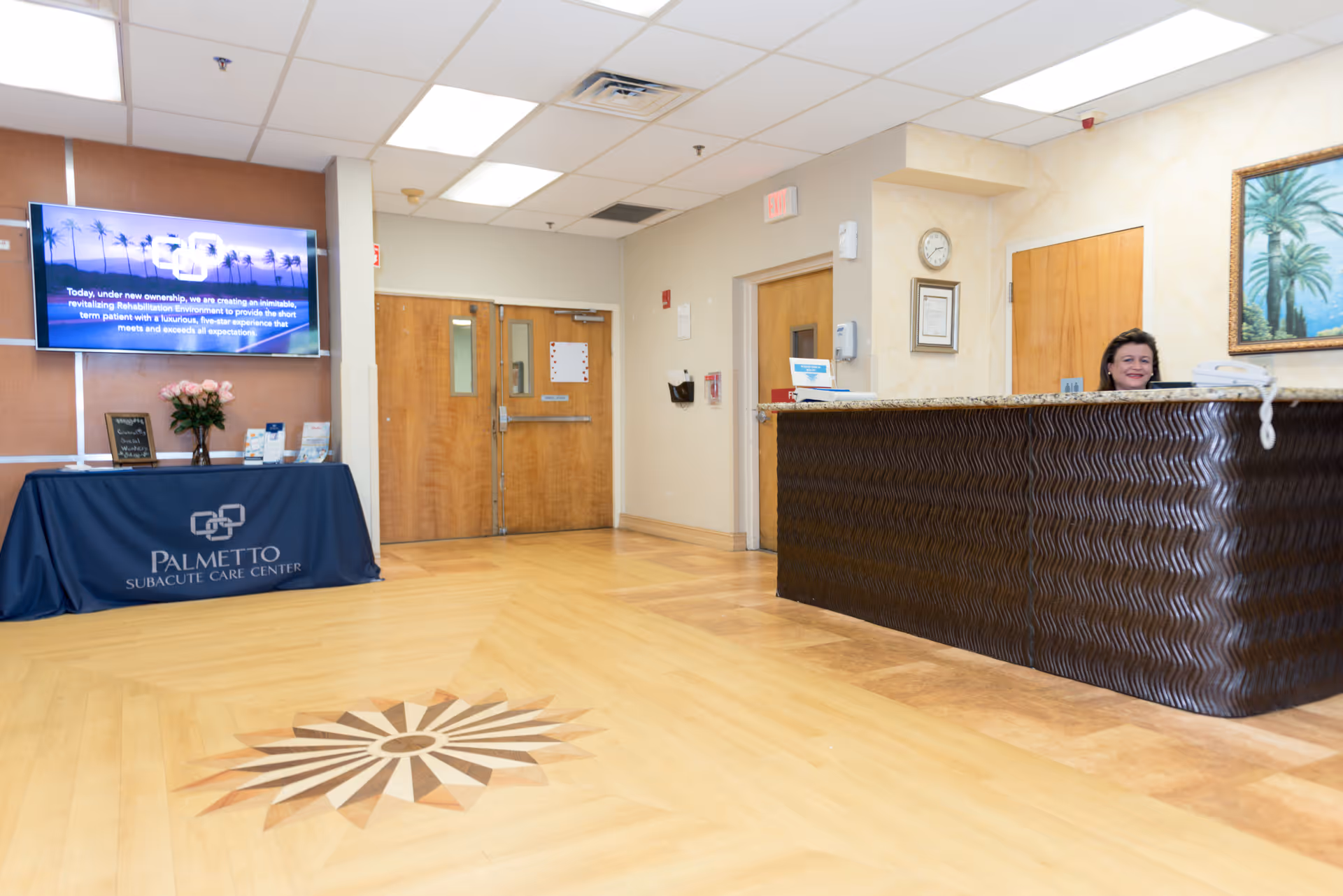 Reception area of Palmetto Subacute Care Center with a front desk where a woman is seated. There is a decorative wooden floor with a sunburst pattern, double wooden doors in the background, a table covered with a blue cloth displaying the facility name and logo, a TV screen mounted on the wall, and a framed painting of palm trees.