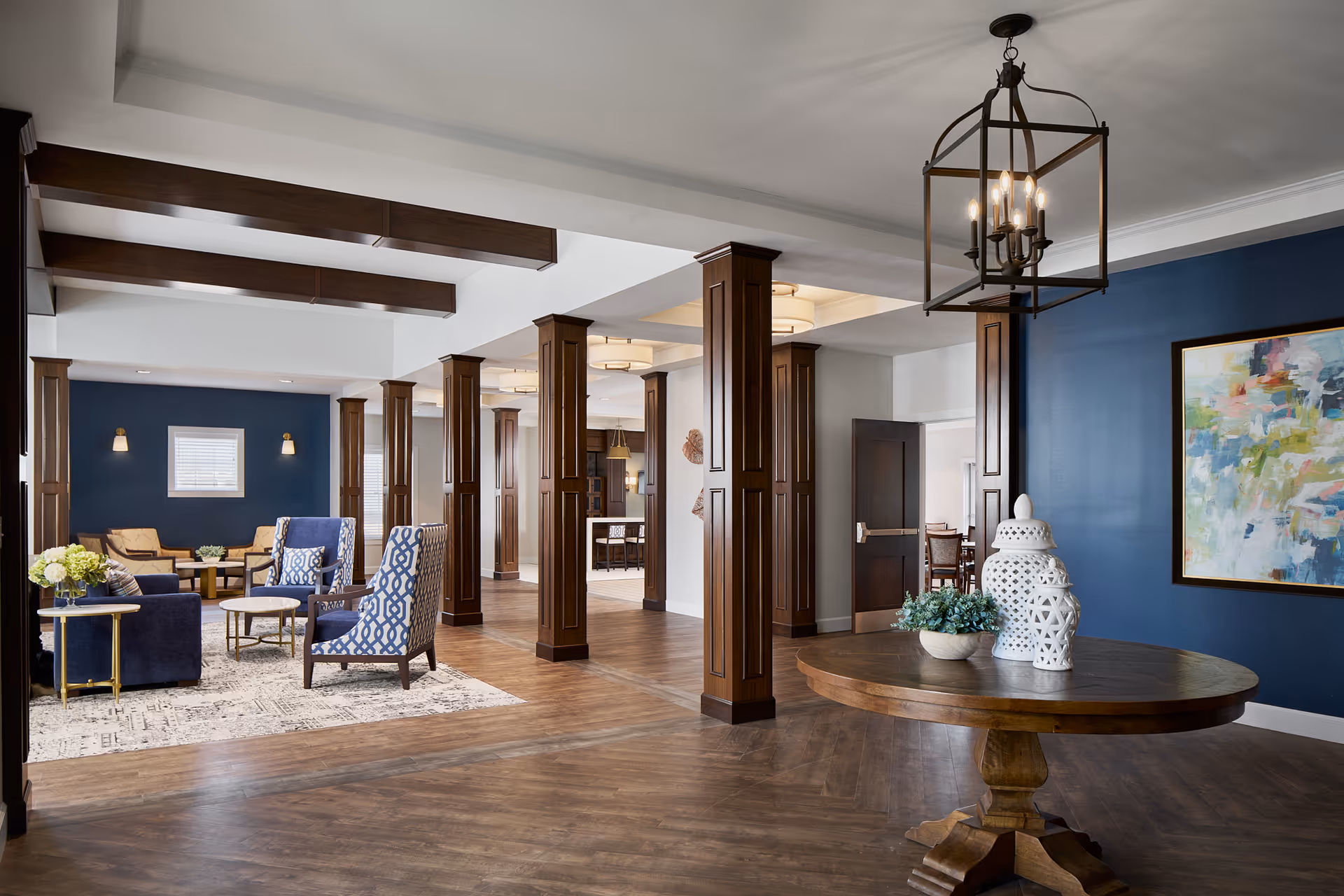 A spacious and elegant common area with wooden floors and dark wood columns. The room features a round wooden table with decorative white ceramic lanterns and a small plant. There is a seating area with blue and patterned armchairs, a sofa, and a coffee table on a patterned rug. The walls are painted blue and white, with a colorful abstract painting on one wall. The ceiling has exposed wooden beams and modern light fixtures.