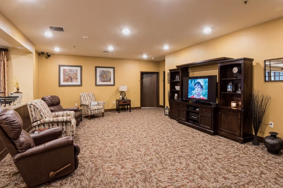 Carpeted communal living room with recliners and armchairs arranged facing a large wooden entertainment center and TV.