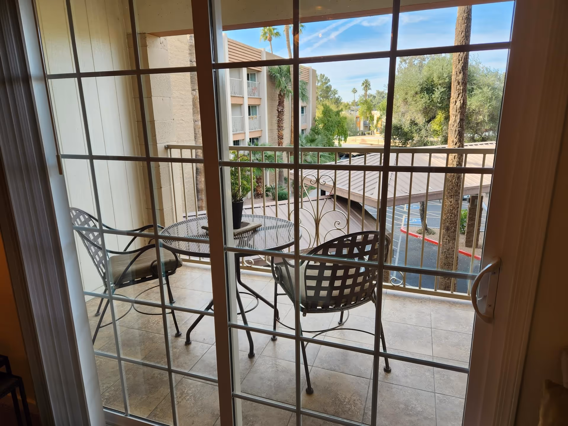 View through a glass sliding door onto a balcony with a round metal table and two metal chairs. The balcony overlooks a parking area, palm trees, and a multi-story building under a blue sky with some clouds.