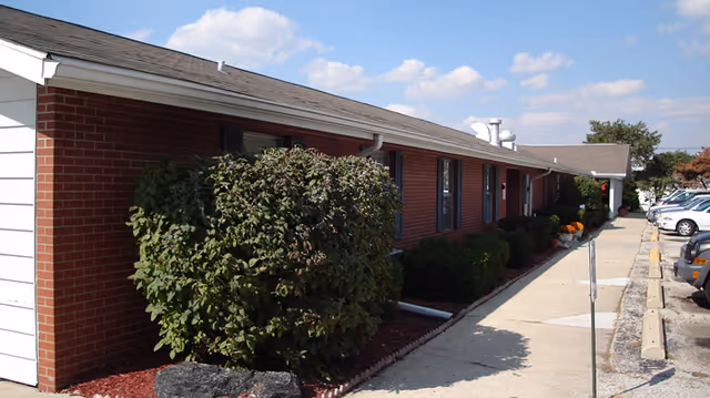 Exterior view of a single-story brick building with a white roof trim, bushes along the side, a sidewalk, and parked cars under a blue sky with some clouds.