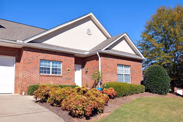 Exterior view of a single-story brick building with a white door, two windows, and a small garden with bushes and shrubs in front. The sky is clear and blue.