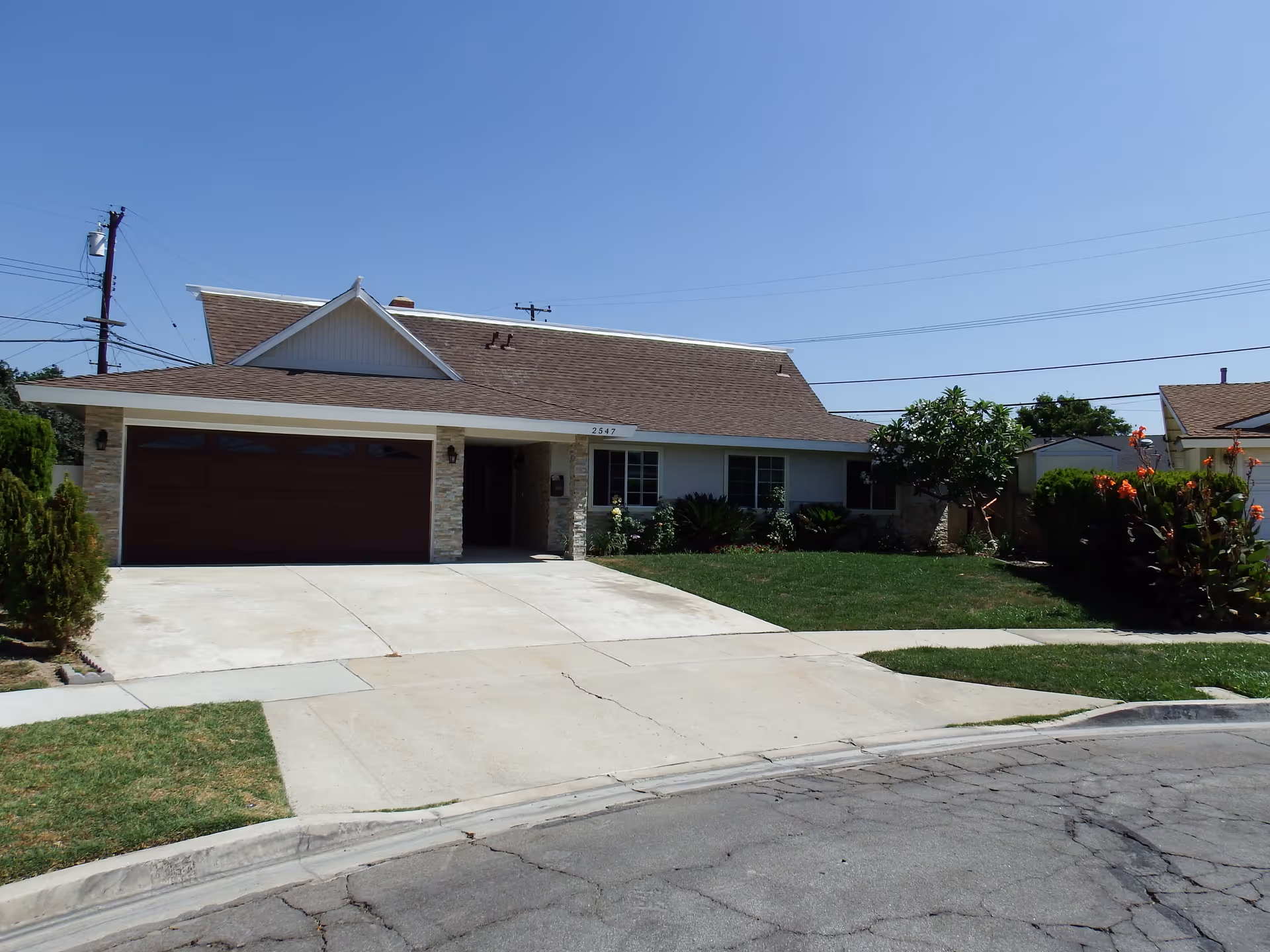 Front exterior view of a single-story residential building with a brown shingle roof, a double garage door, a concrete driveway, and a well-maintained lawn with shrubs and flowering plants under a clear blue sky.