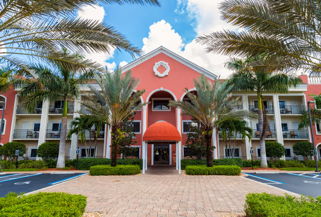 Front exterior view of a three-story senior living facility building with a red facade, white columns, and a red awning over the entrance. The building is surrounded by palm trees and neatly trimmed bushes, with a paved walkway leading to the entrance and parking spaces on either side.
