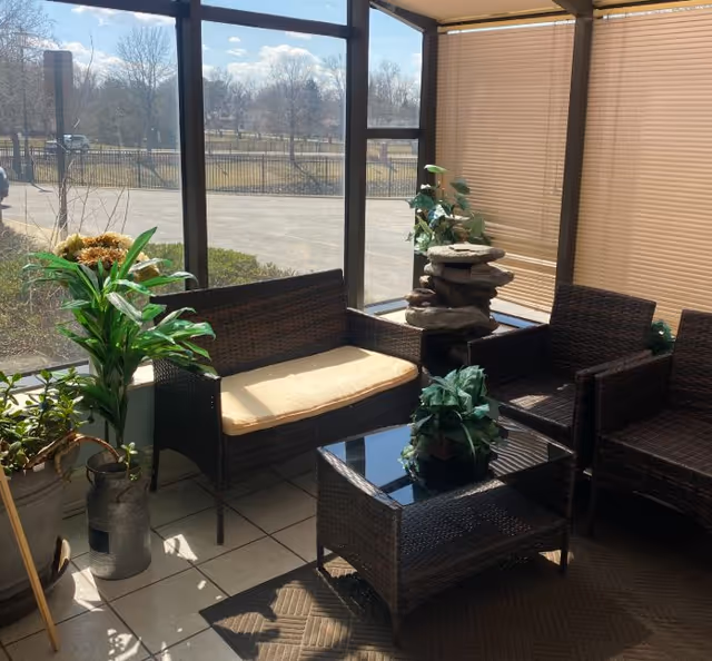Sunlit indoor seating area with wicker furniture including a cushioned loveseat, two chairs, and a glass-top coffee table. Several potted plants and a decorative stone fountain are placed around the seating area. Large windows with beige blinds allow natural light to fill the space and provide a view of an outdoor parking lot and trees.