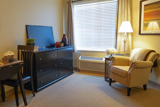 A cozy living room area with a beige armchair next to a small round side table holding a lamp and a vase of flowers. A large window with blinds and curtains lets in natural light. Across from the chair is a black dresser with decorative items and a flat-screen TV on top. A wooden chair and small table are partially visible on the left side.