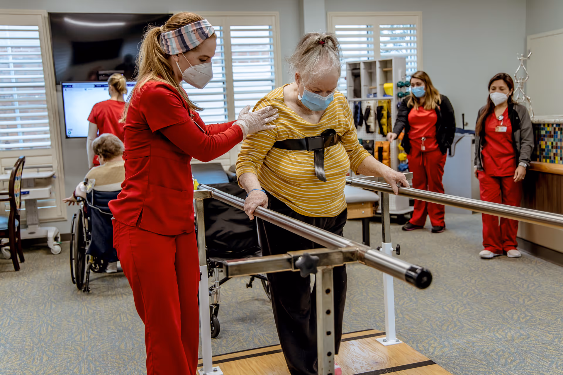 A healthcare worker in red scrubs and a face mask assists an elderly woman wearing a yellow striped shirt and face mask as she practices walking between parallel bars in a rehabilitation center. In the background, two other healthcare workers in red scrubs and face masks observe, and another elderly person is seated in a wheelchair near a window.