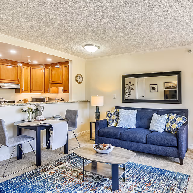 A cozy living room and dining area in a senior living facility. The living room features a blue sofa with patterned and solid pillows, a wooden coffee table with a decorative bowl, and a large mirror on the wall. Adjacent to the living room is a small dining area with a round table set for two, two gray chairs, and a kitchen with wooden cabinets and granite countertops in the background.