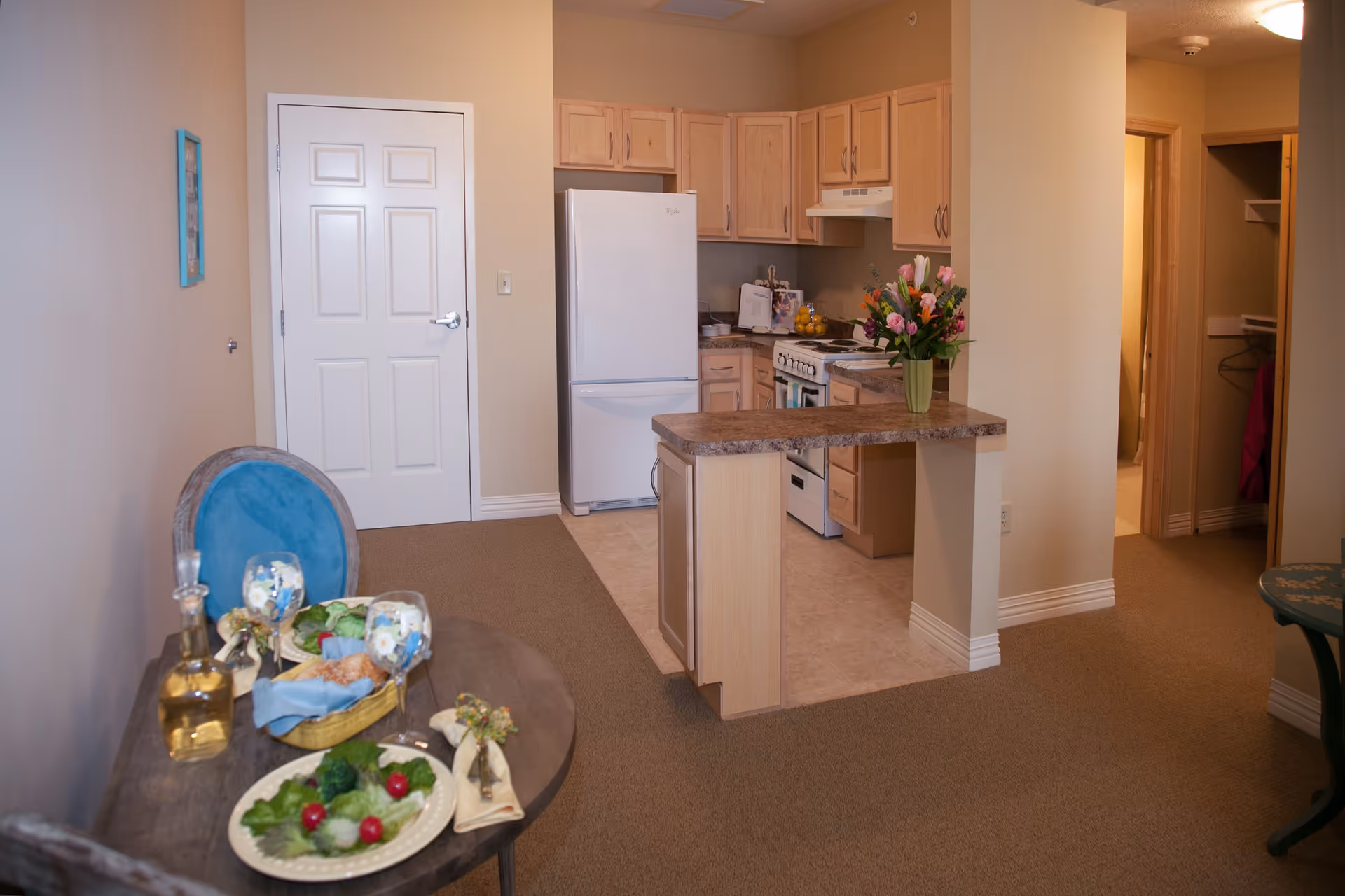 Interior view of a senior living facility apartment showing a small kitchen with light wood cabinets, a white refrigerator, stove, and a countertop with a vase of flowers. In the foreground, there is a round table set with plates of salad, glasses, and a bottle. The room has beige walls and carpeted floors, with a door and an open closet visible in the background.