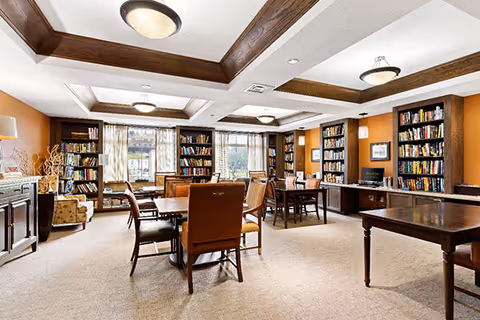A spacious and well-lit library or reading room with multiple wooden tables and chairs arranged throughout. The room features built-in bookshelves filled with books along the walls, large windows with curtains allowing natural light to enter, and ceiling lights with decorative wooden beams. There is a computer workstation on one side and comfortable seating areas near the windows.