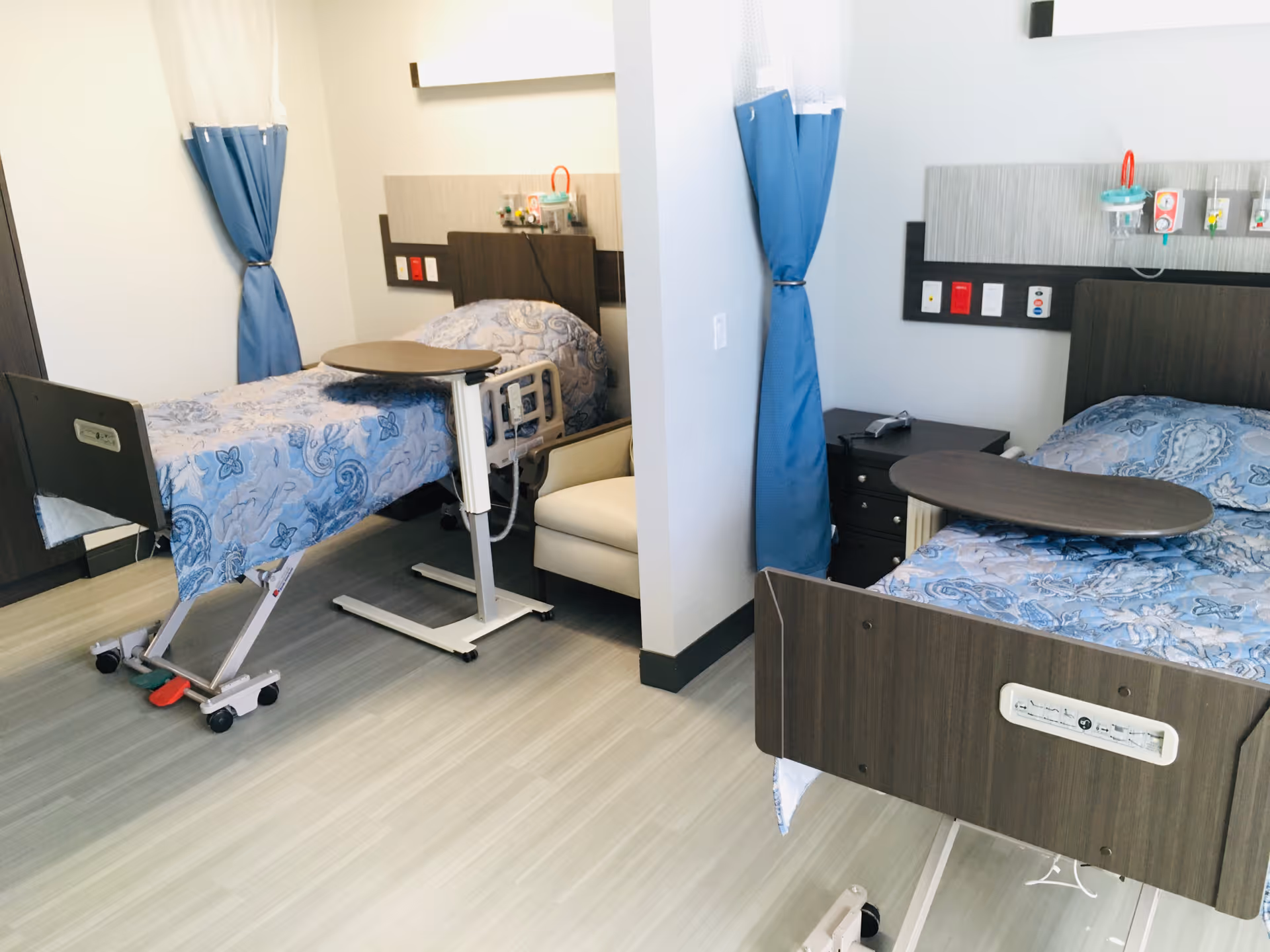 Two hospital-style beds with blue patterned bedding, overbed tables, and privacy curtains in a shared nursing facility room.
