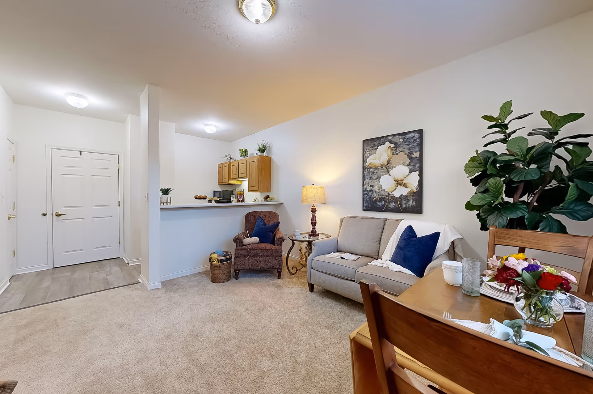 A cozy living area in an assisted living facility featuring a beige sofa with blue pillows and a white throw, a patterned armchair with a blue pillow, a small round side table with a lamp, and a dining table set with flowers and tableware. The room has light-colored walls, carpeted flooring, and a small kitchen area with wooden cabinets and a counter in the background.