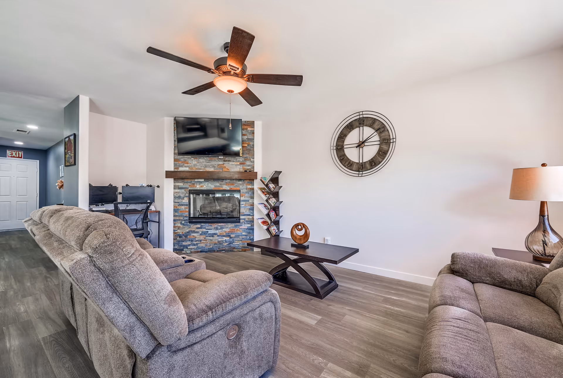 A cozy living room with two gray reclining sofas facing a stone fireplace with a mounted flat-screen TV above it. A modern dark wood coffee table with a decorative piece sits in the center. A large wall clock is mounted on the white wall, and a table lamp with a glass base is on a side table next to one sofa. In the background, there is a hallway with an exit sign and a small desk with two computer monitors and chairs.
