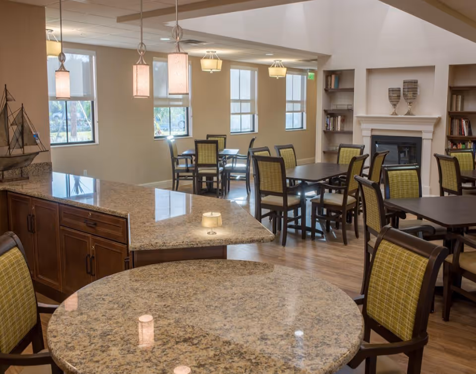 A well-lit dining area in a senior living facility featuring multiple tables and chairs with green plaid upholstery. The room has large windows with white blinds, a fireplace with decorative items on the mantel, built-in bookshelves, and hanging pendant lights above a granite countertop.