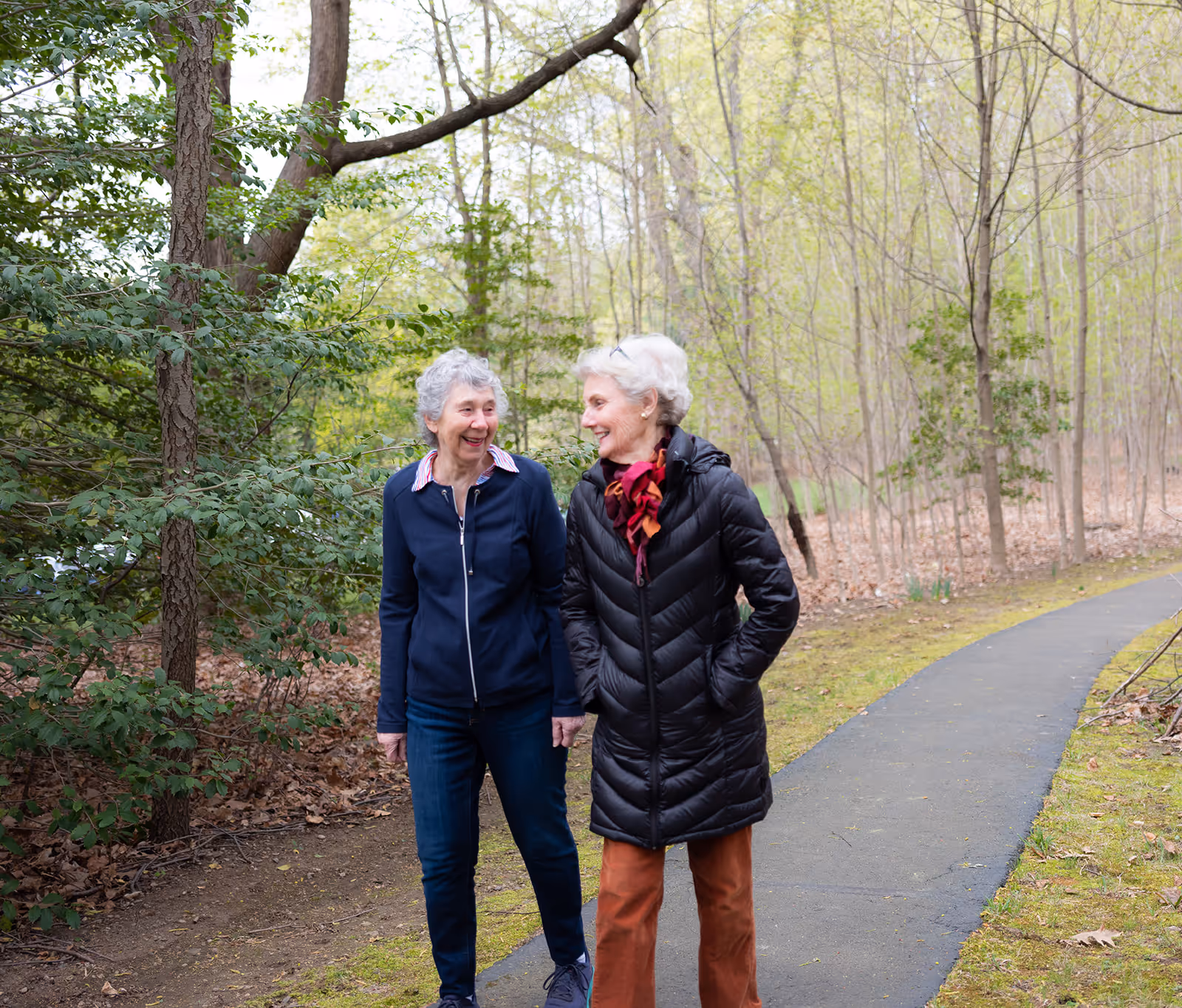 Two elderly women walking and smiling together on a paved path in a wooded outdoor area with trees and greenery around them.