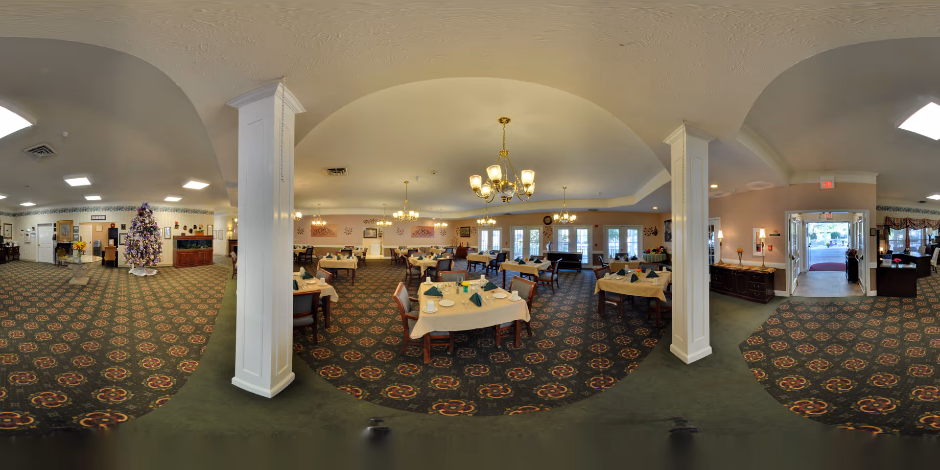 A spacious dining room in a senior living facility with multiple tables covered with white tablecloths, set with plates, cups, and folded napkins. The room features patterned carpet, chandeliers hanging from the ceiling, large windows letting in natural light, and decorative wall art. There is a Christmas tree decorated with purple and white ornaments near the entrance area.