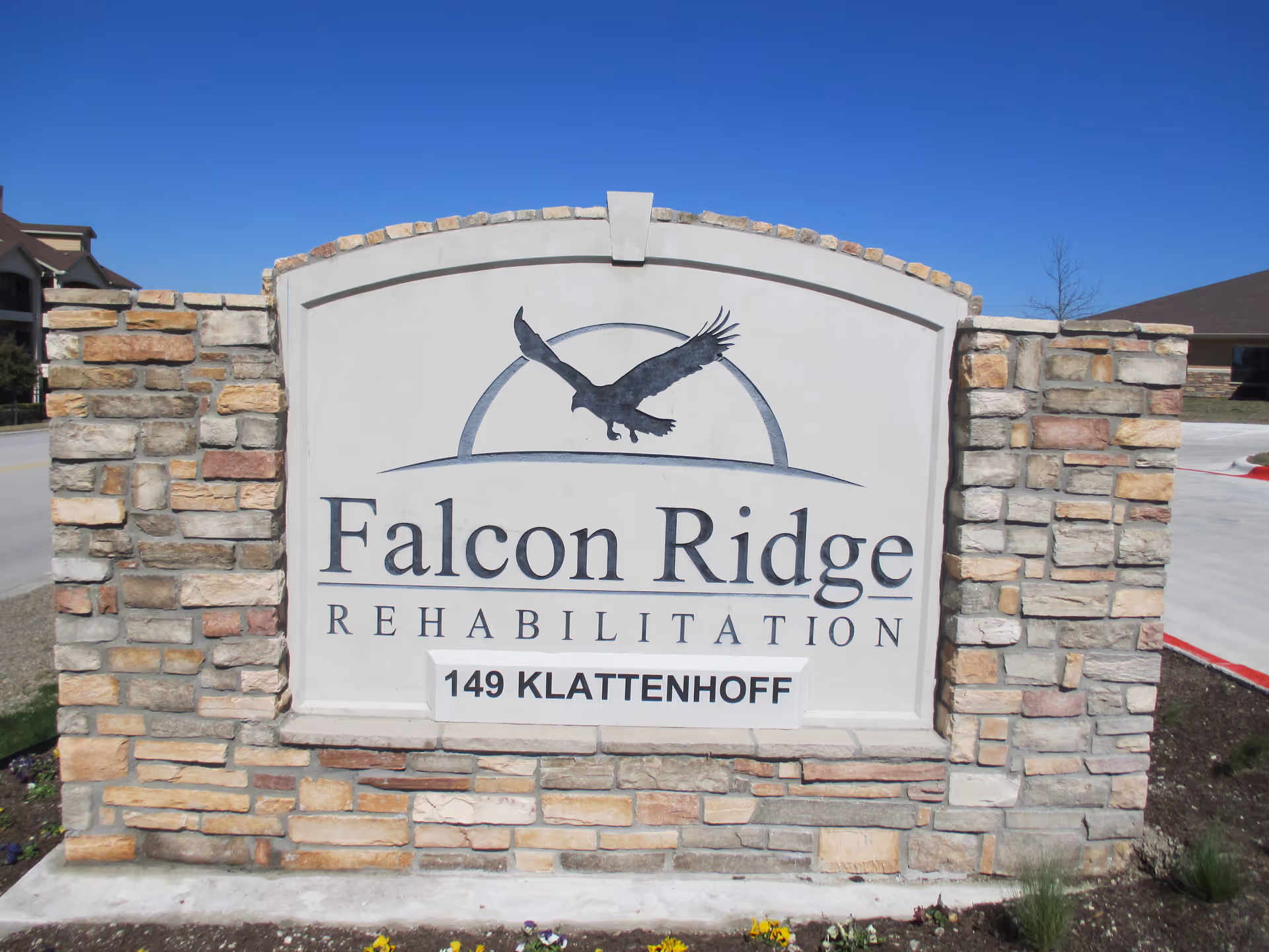 Stone sign for Falcon Ridge Rehabilitation with an image of a flying falcon above the facility name and the address 149 Klattenhoff below, set against a clear blue sky.