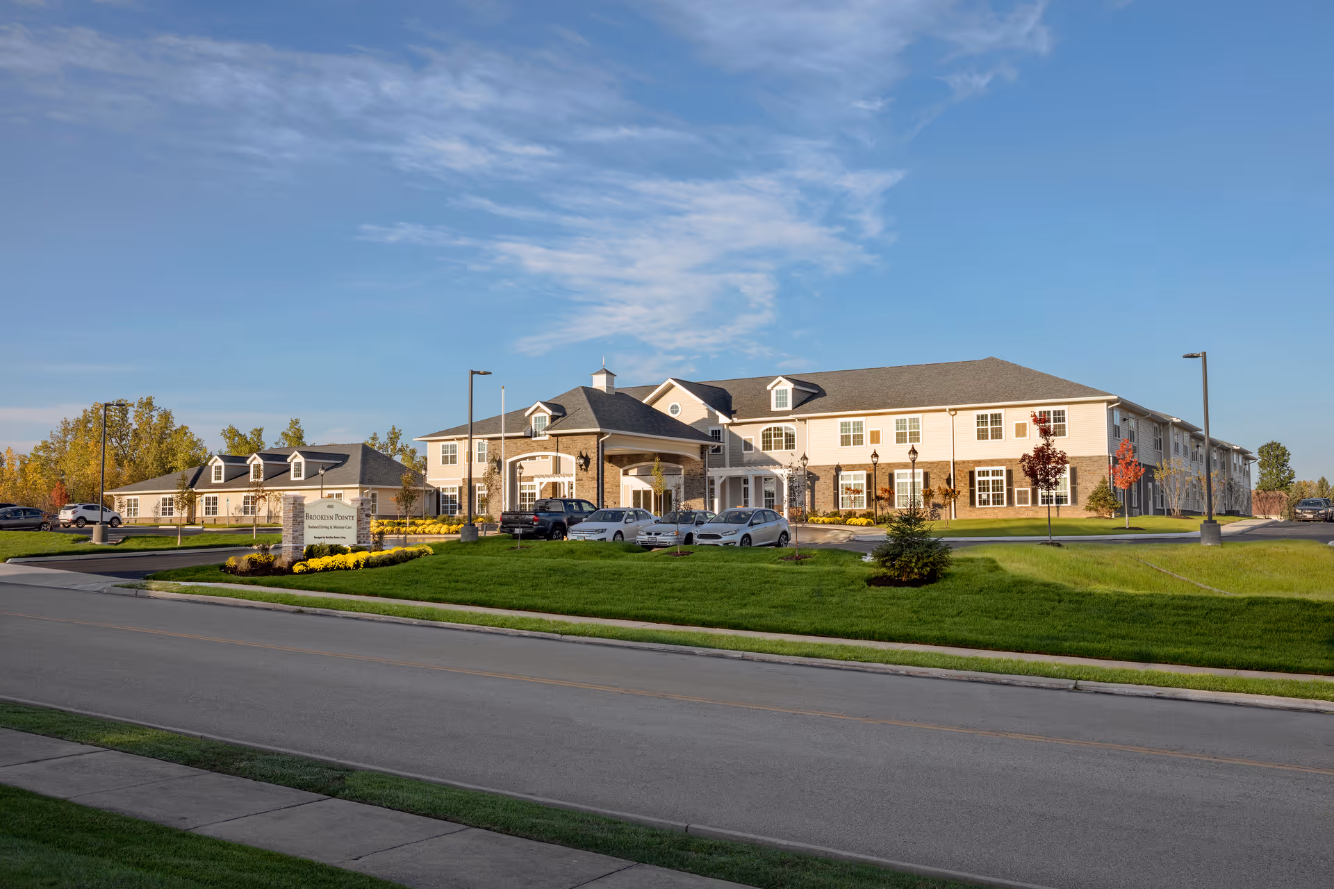 Exterior view of Brooklyn Pointe Assisted Living & Memory Care facility showing a large two-story building with multiple windows, a covered entrance with parked cars, well-maintained green lawns, and a clear blue sky.