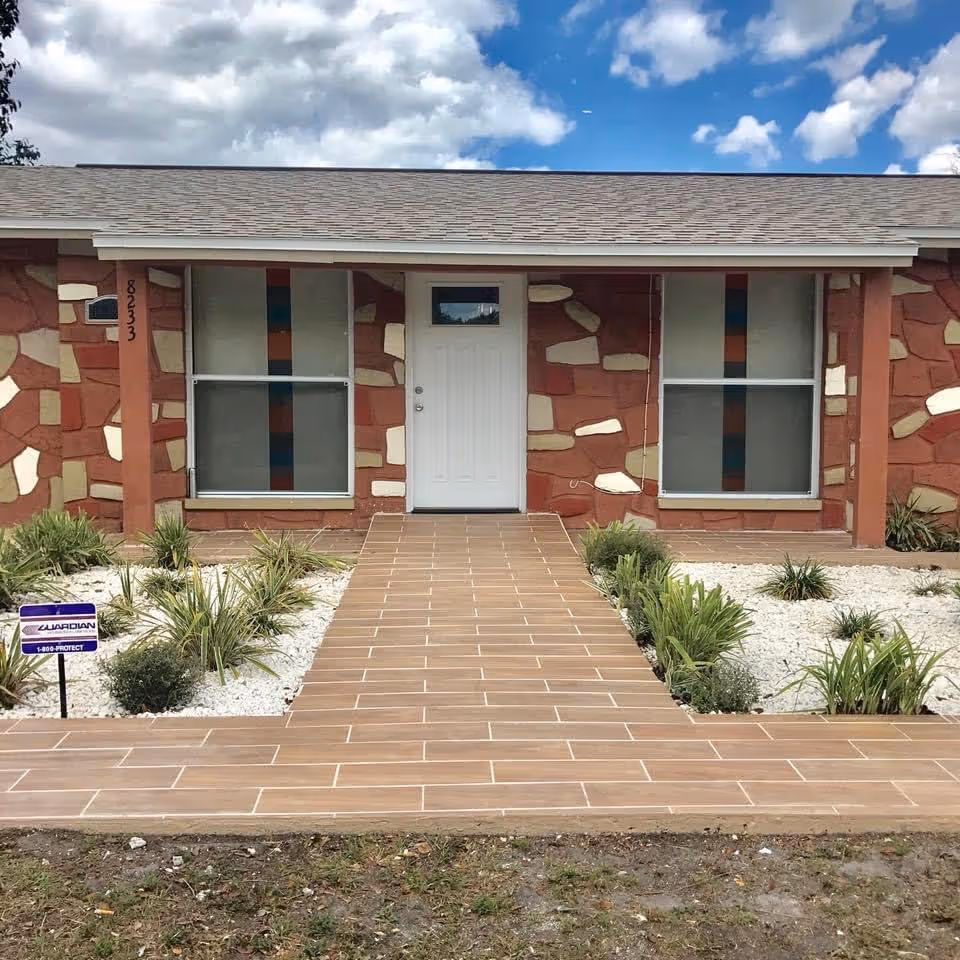 Front exterior view of a single-story building with a tiled walkway leading to a white door. The building has a unique stone-patterned facade with red, beige, and brown colors. Two large windows with vertical colored stripes flank the door. There are small landscaped garden beds with green plants and white gravel on either side of the walkway. The sky is partly cloudy.