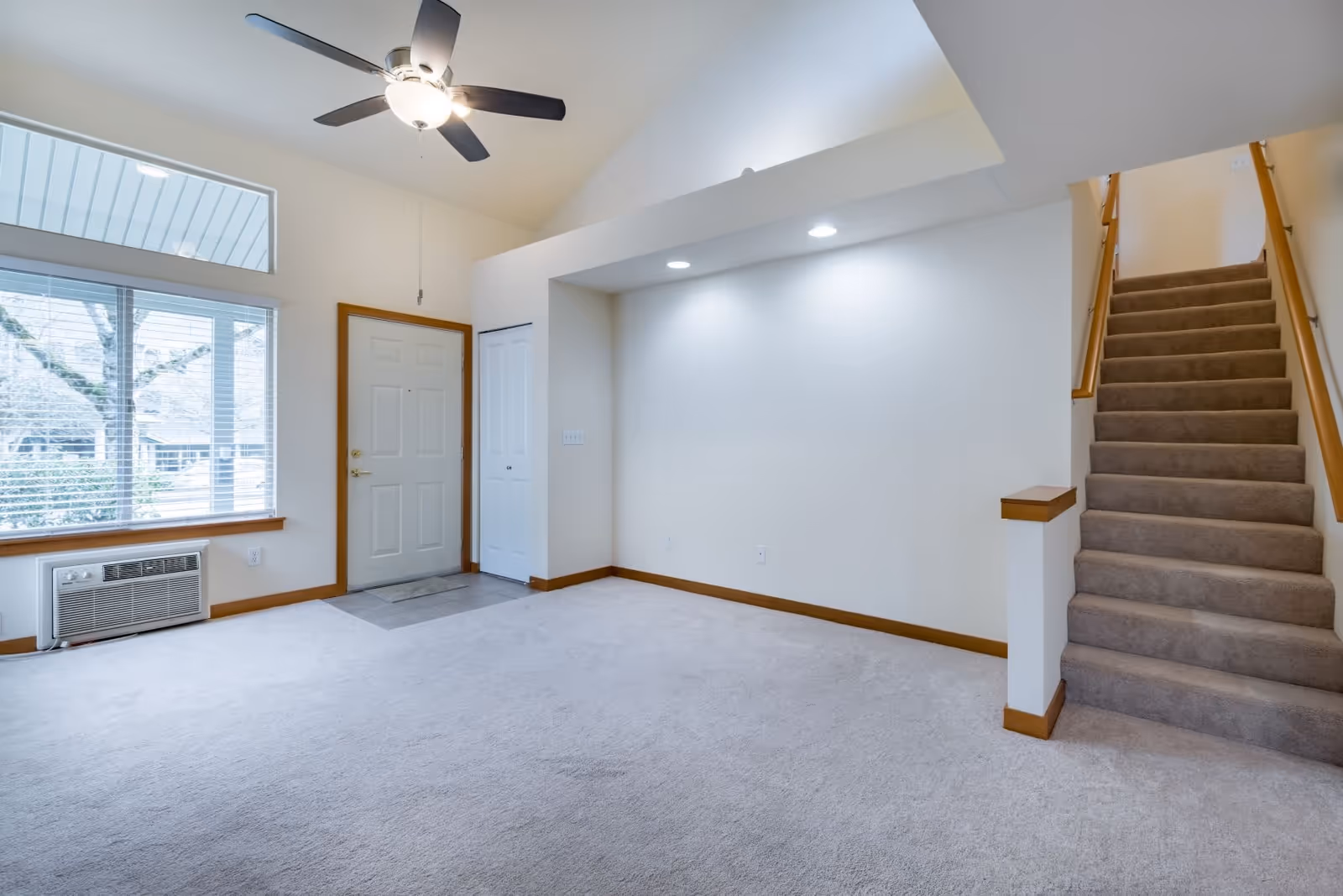 Empty interior room with beige carpet, white walls, a ceiling fan with light, a window with blinds, a white front door with a small closet next to it, and a carpeted staircase with wooden handrails leading upstairs.