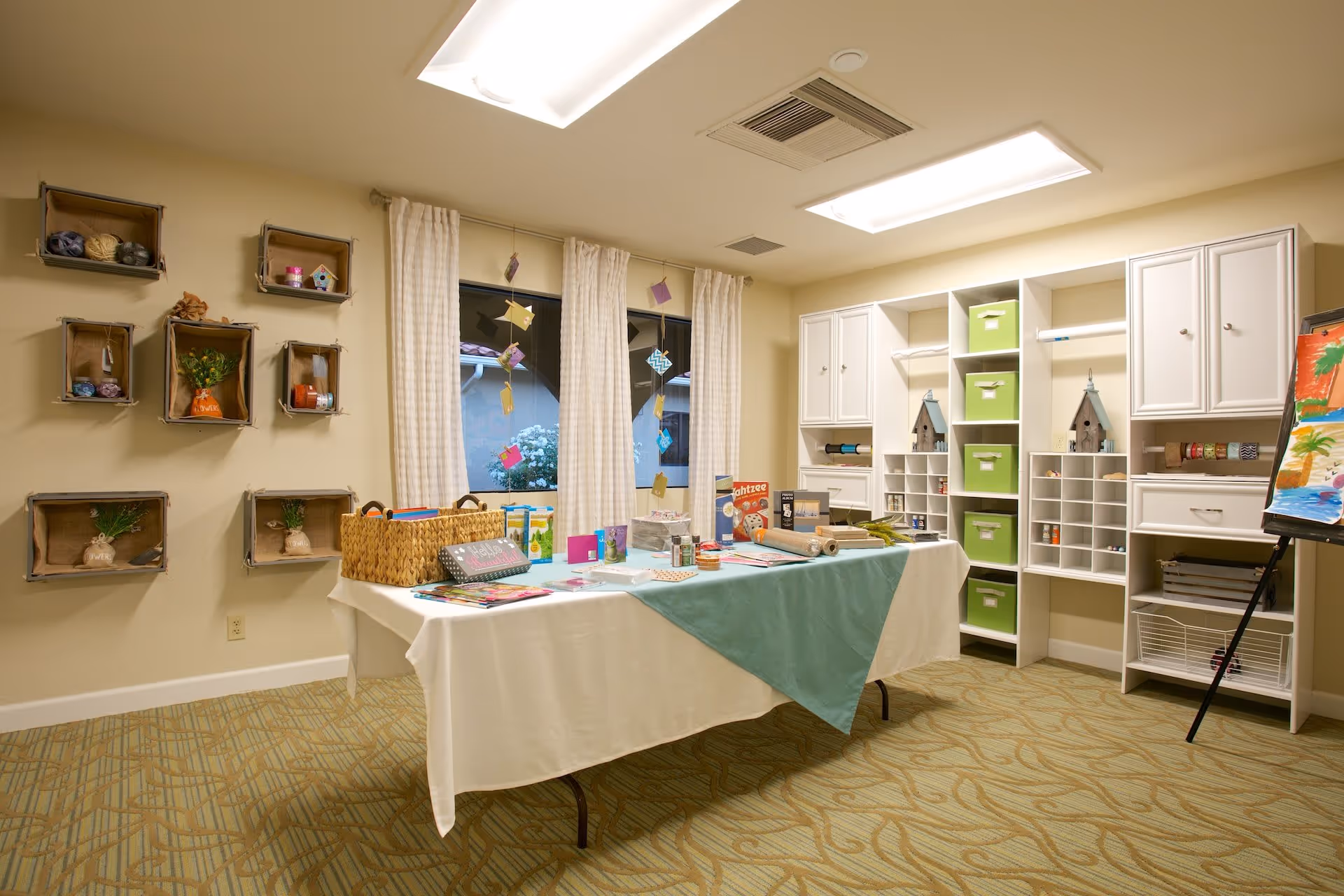 A well-lit craft room with a table covered by a white and teal tablecloth, displaying various craft supplies and games. The room has beige walls with decorative shelves holding yarn and small items, a window with white curtains, and white built-in cabinets and cubbies with green storage bins. An easel with colorful paintings is visible on the right side.