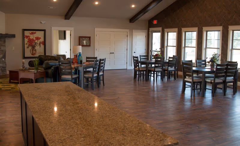A spacious dining area with several wooden tables and chairs arranged near large windows letting in natural light. The room features wooden flooring, a granite countertop in the foreground, and a cozy seating area with a sofa and side tables. The walls have decorative artwork and the ceiling has exposed wooden beams.