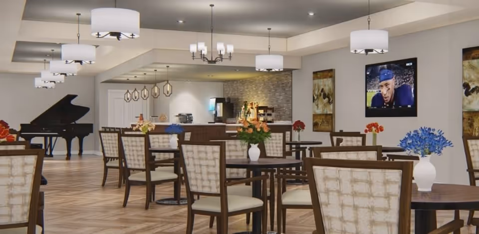 Spacious senior community dining/lounge with multiple tables and chairs, floral centerpieces, a grand piano and a wall-mounted TV.